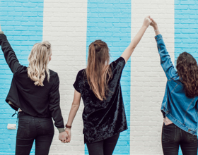 Tres mujeres tomadas de la mano frente a una pared colorida.