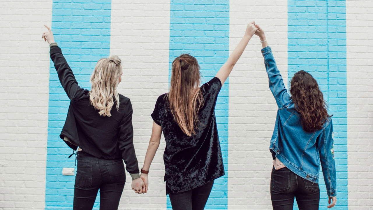 Tres mujeres tomadas de la mano frente a una pared colorida.