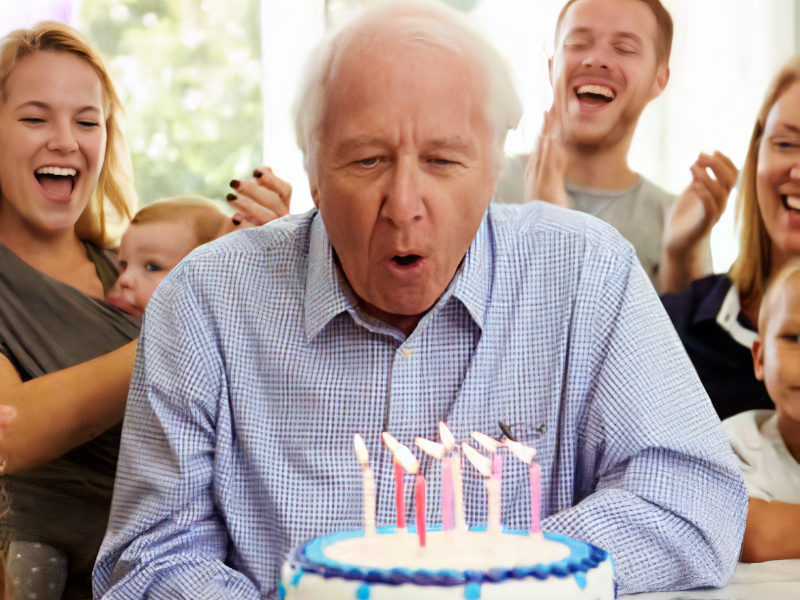 Photo of an elderly man at a birthday party blowing out the candles on a birthday cake.